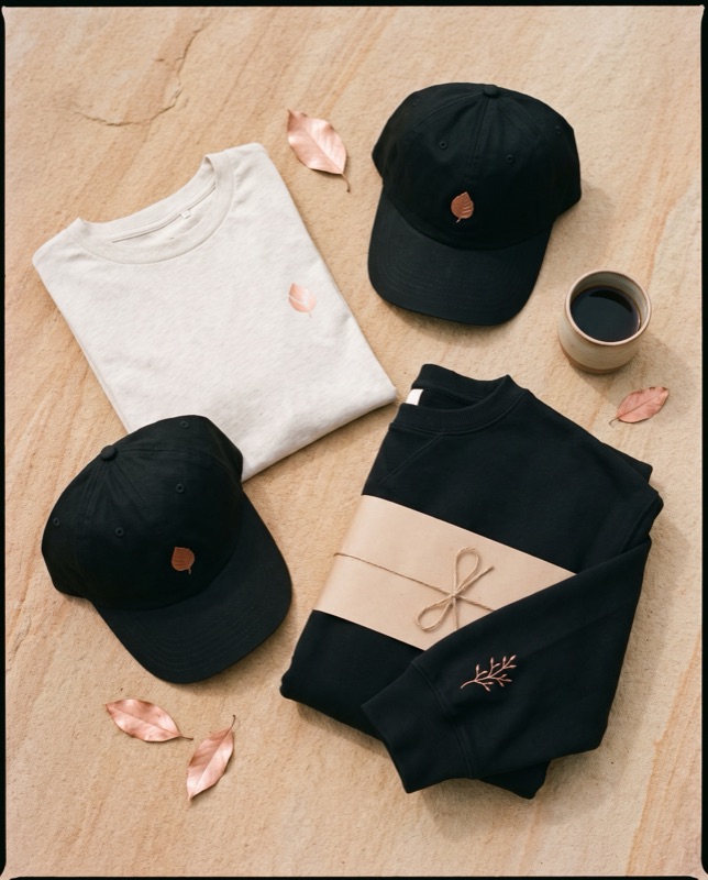 Flat lay of a cream tee, two black caps with a copper leaf and a black crewneck with a copper sprig on the sleeve, beside a cup of coffee