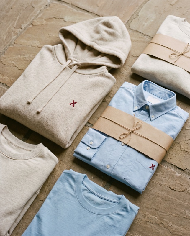 Flat lay of an oatmeal hoodie with an oxblood serif mark, a light-blue oxford shirt, a light-blue tee and oatmeal tees, tied with paper bands