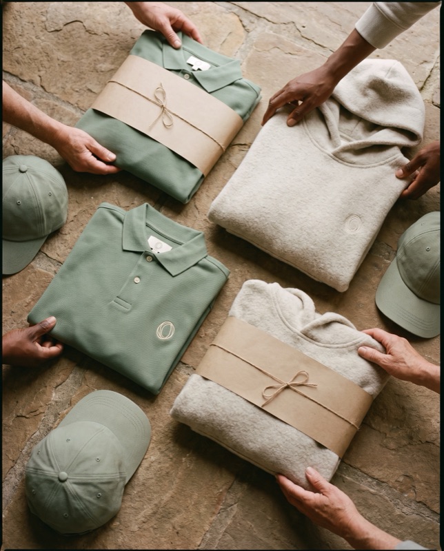Flat lay of sage piqué polos with a cream embroidered mark, cream hoodies and sage caps, tied with paper bands