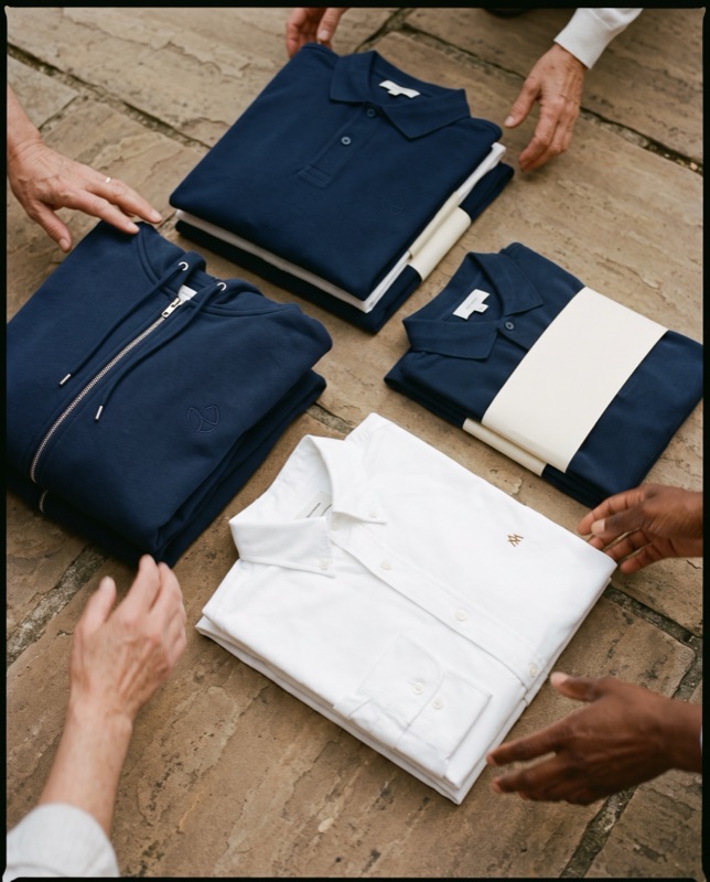 Flat lay of navy piqué polos, a navy zip hoodie and a white oxford shirt with a small brass monogram, on stone