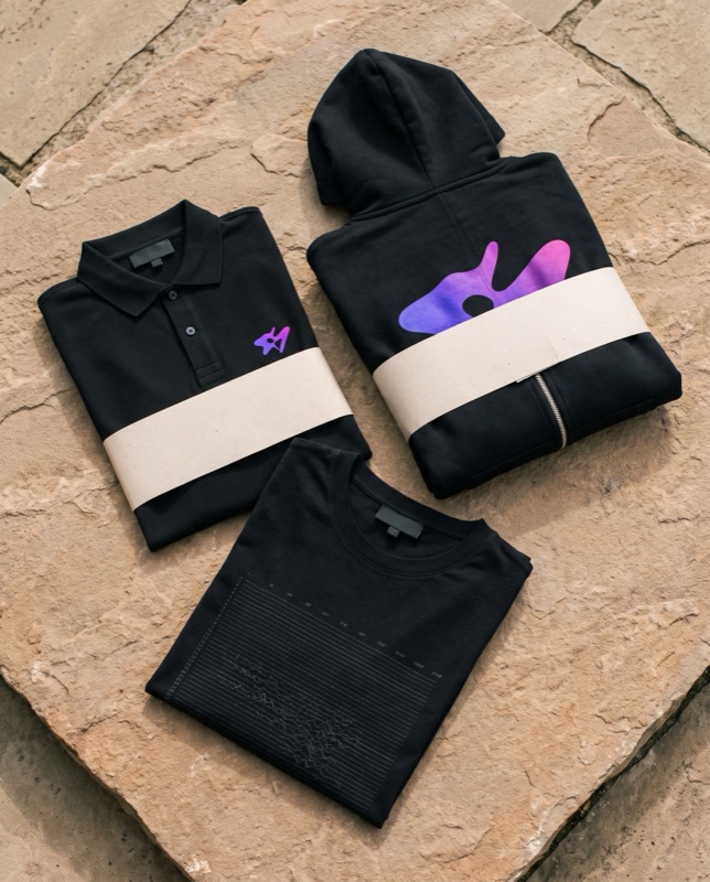 Flat lay of a black polo and a black zip hoodie with electric violet marks and a black tee with a tonal print, on stone