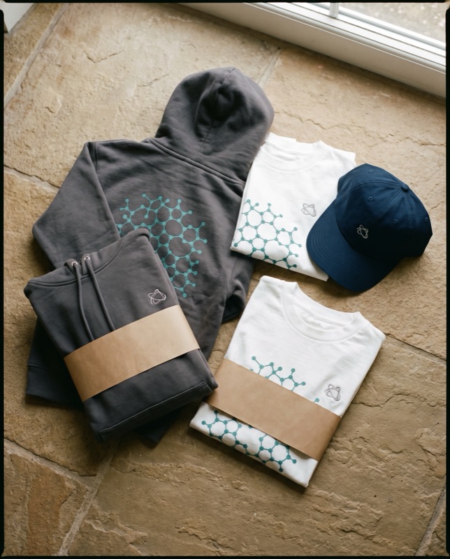Flat lay of charcoal hoodies and white tees printed with a teal molecular lattice and a navy cap, on stone