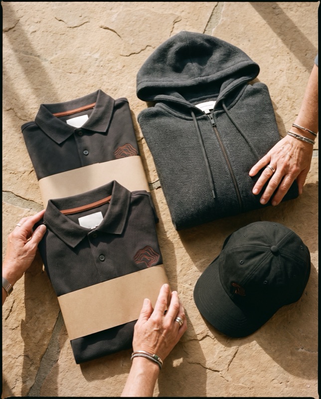 Flat lay of charcoal polos with a rust embroidered topographic mark, a charcoal zip hoodie and a black cap, on stone