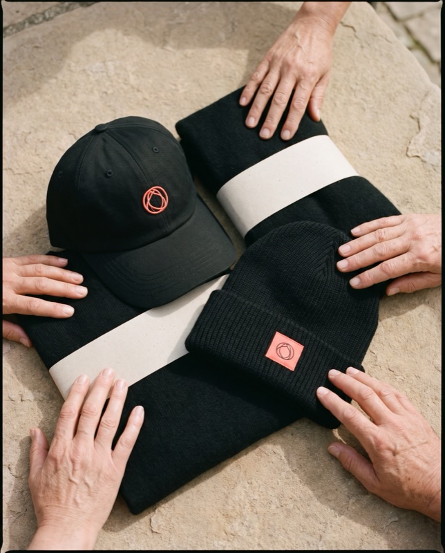 Flat lay of a black cap with a coral embroidered mark and black cuffed beanies with a coral label patch